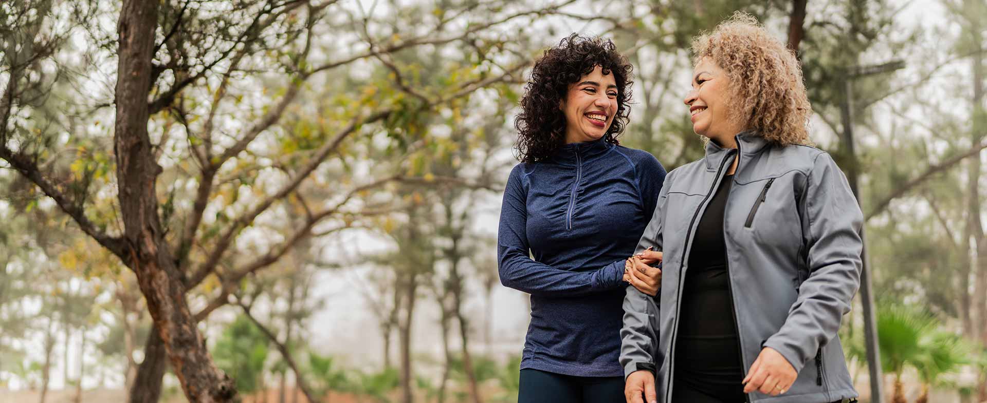 Two women walking and smiling at each other in a tree-lined park path. They are dressed in casual athletic wear. The background features trees and an overcast sky. 10k to 7k steps