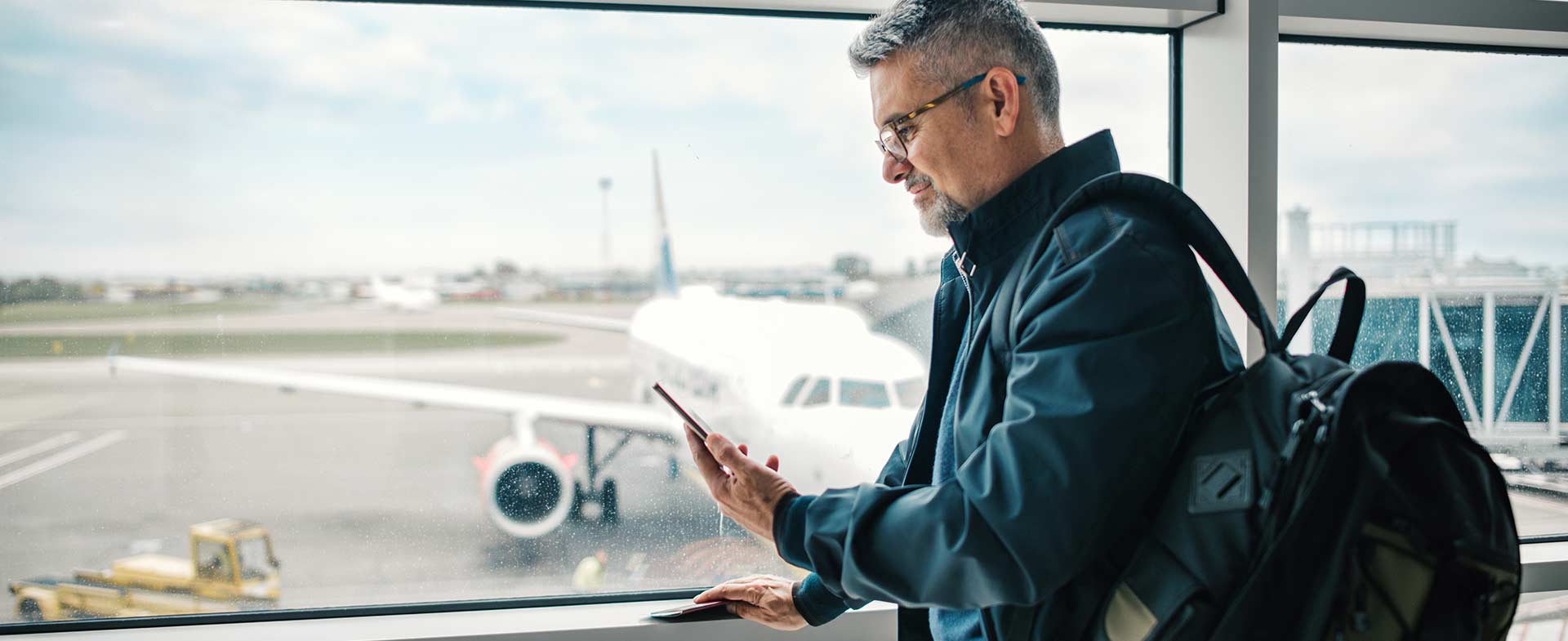 Middle aged man with glasses wearing a navy jacket looking at cell phone in front of an airport window flying and heart heart