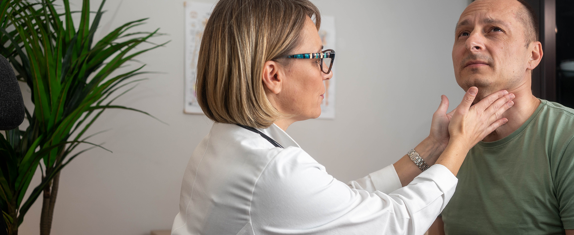 Female doctor with short blonde hair wearing a white coat and checking for thyroid nodules on male patient's neck. Male patient is wearing a green short sleeved t-shirt. doctor checking thyroid nodules