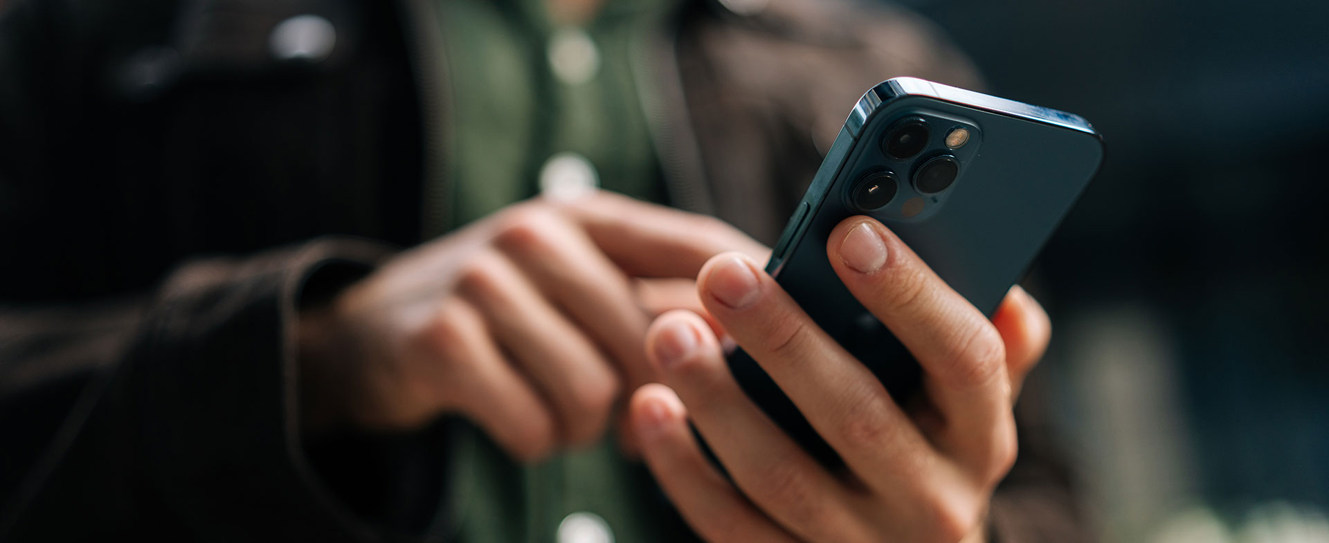 A person is holding and using an iPhone outdoors, wearing a dark jacket and a green shirt. The background is slightly blurred. dystextia