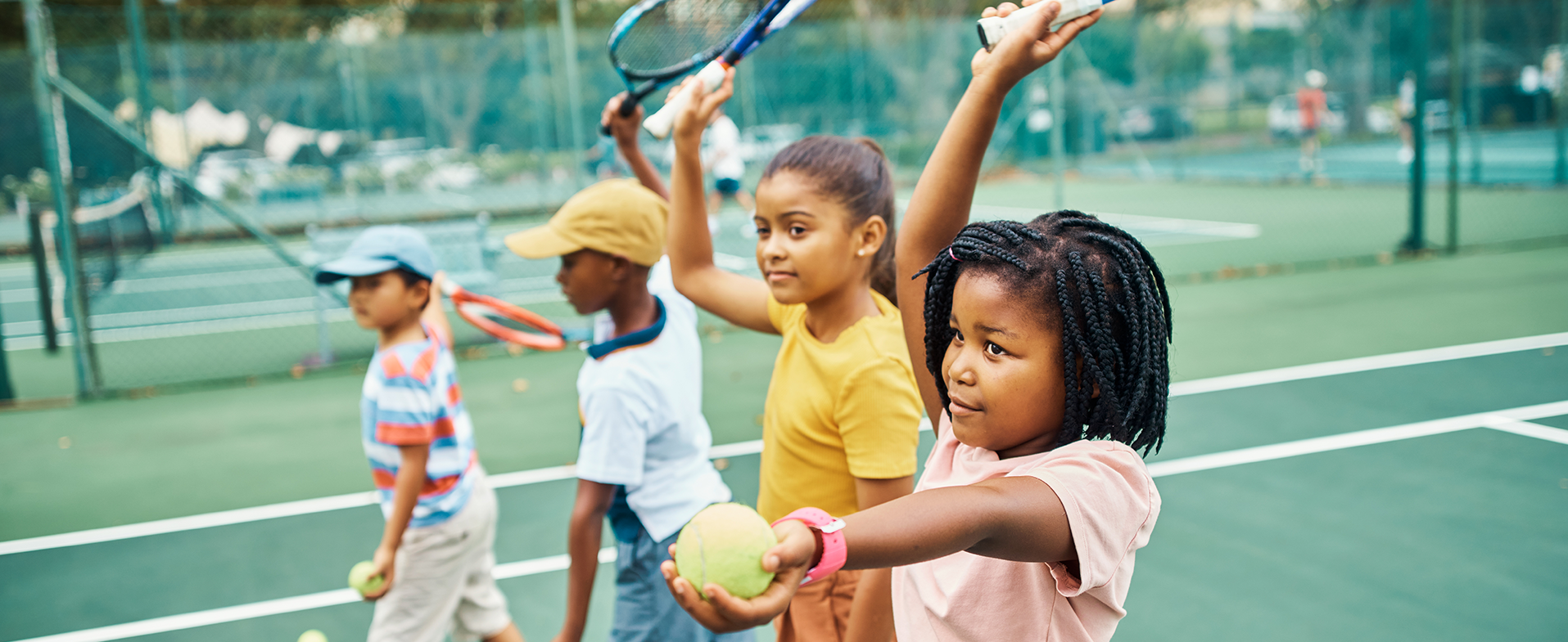Two girls wearing t-shirts - one light peach and one yellow - and two boys wearing t-shirts - one white with a navy trim and one blue, red and white striped. They are all outside on a tennis court playing tennis. dummy desktop Image