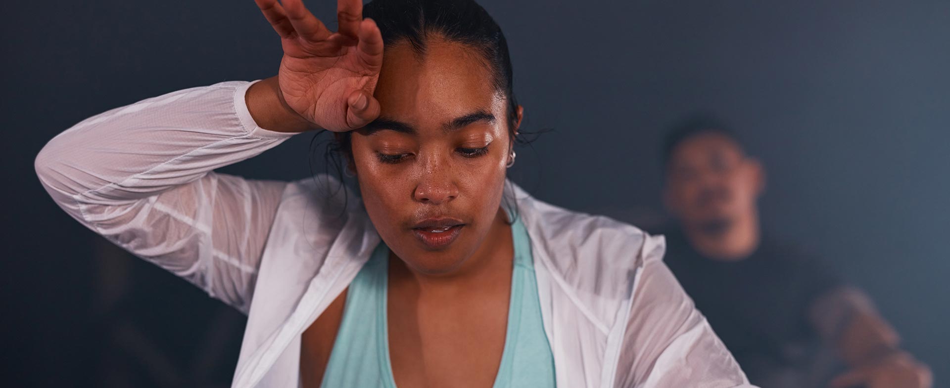 A woman wearing a light blue top and white jacket wipes her forehead after working out energetically. Another person is visible in the background, slightly out of focus. The setting appears dimly lit, creating a dynamic atmosphere. hot workouts