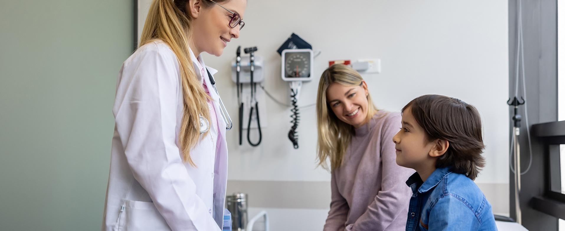 Little boy wearing blue button down shirt at pediatrician appointment with his mom who has long blonde hair and is wearing a lilac sweater. The pediatrician is wearing a white coat with glasses and has her hair in a ponytail. dummy desktop Image