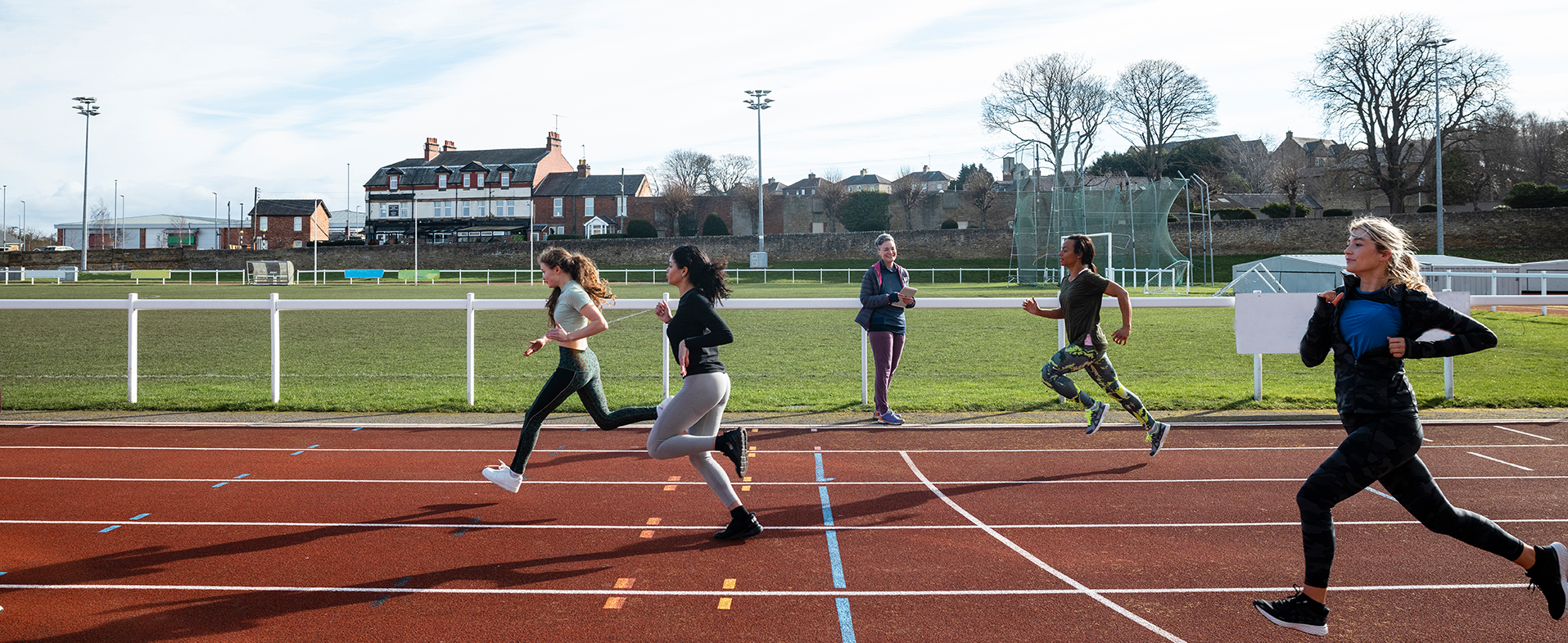 High schoolers wearing leggings, athletic tops and sneakers and running on a track outside. One person is standing alongside the track watching. dummy desktop Image