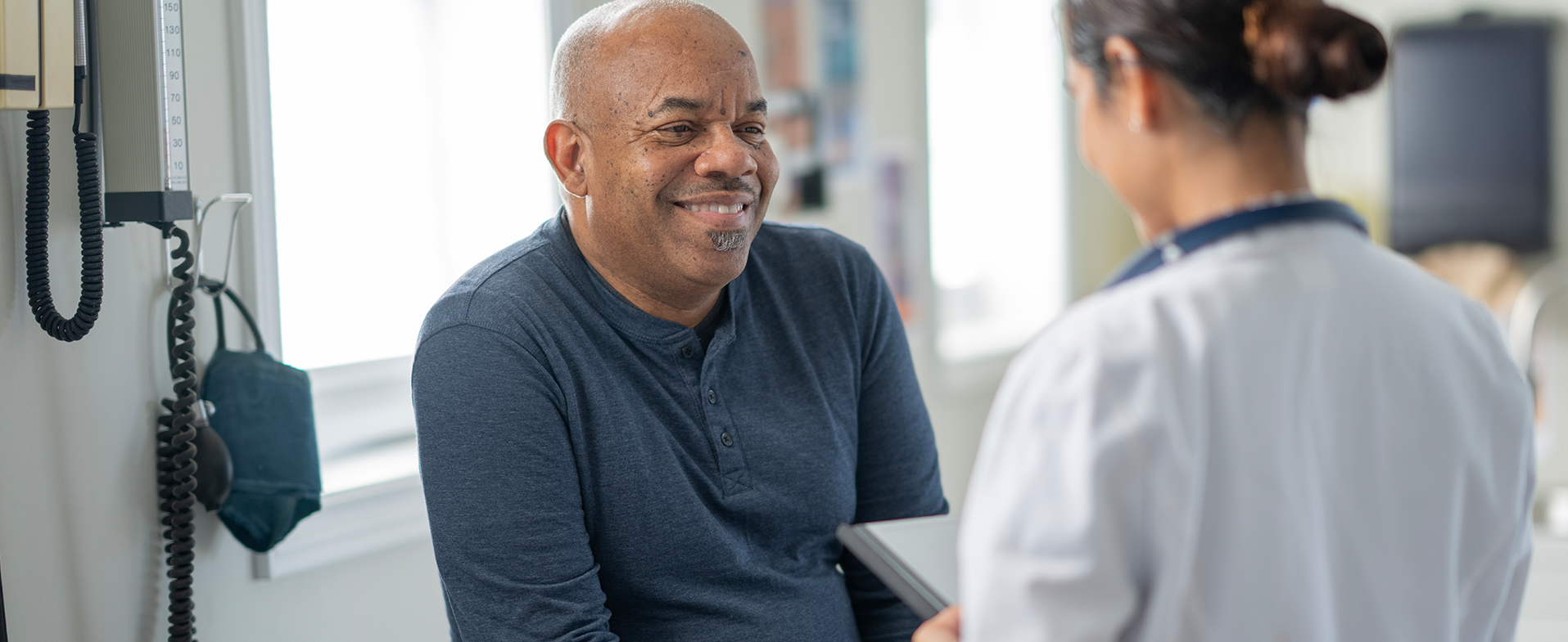 Bald man wearing navy long-sleeved henley shirt smiling and talking to doctor. Her hair is in a low bun and she is wearing a white coat. doctor talking to patient