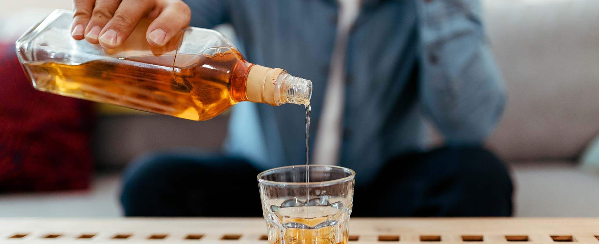 A man with a white beard pours a dark liquor such as whiskey or rum from a bottle into a glass on a wooden table. The background shows a sofa with cushions. heavy drinking and stroke
