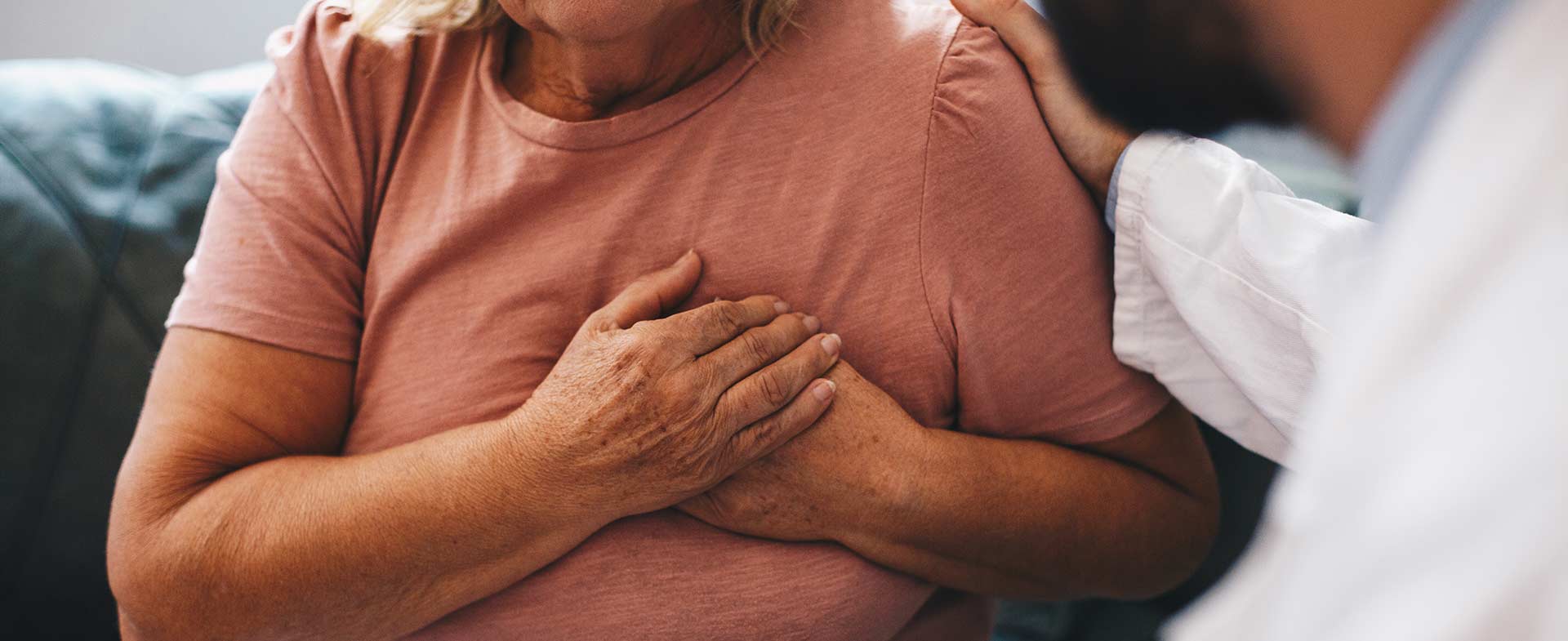 A woman in a pink shirt is seated on a couch with her hands over her heart. A male doctor sits opposite of her with a hand on her shoulder in comfort. pain after heart attack