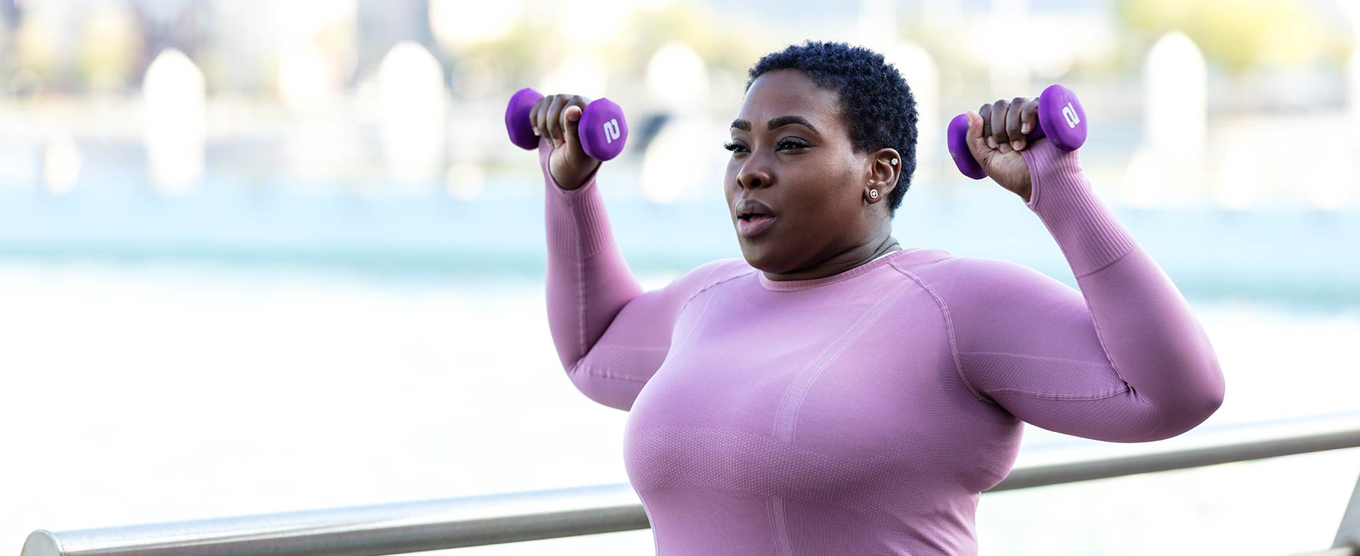 A Black woman in a purple long-sleeve shirt exercises outdoors with lightweight purple dumbbells. The background includes a blurred view of water and buildings. reversing type 2 diabetes