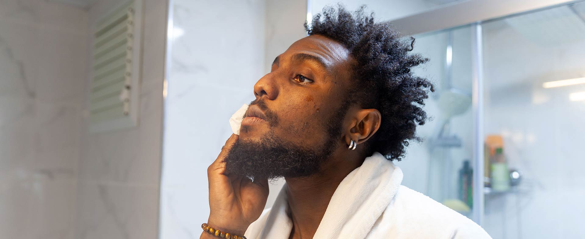 A man with dark skin in a white bathrobe is standing in a bathroom, cleansing their face. The background shows a tiled wall and a glass shower door. skin conditions in dark skin