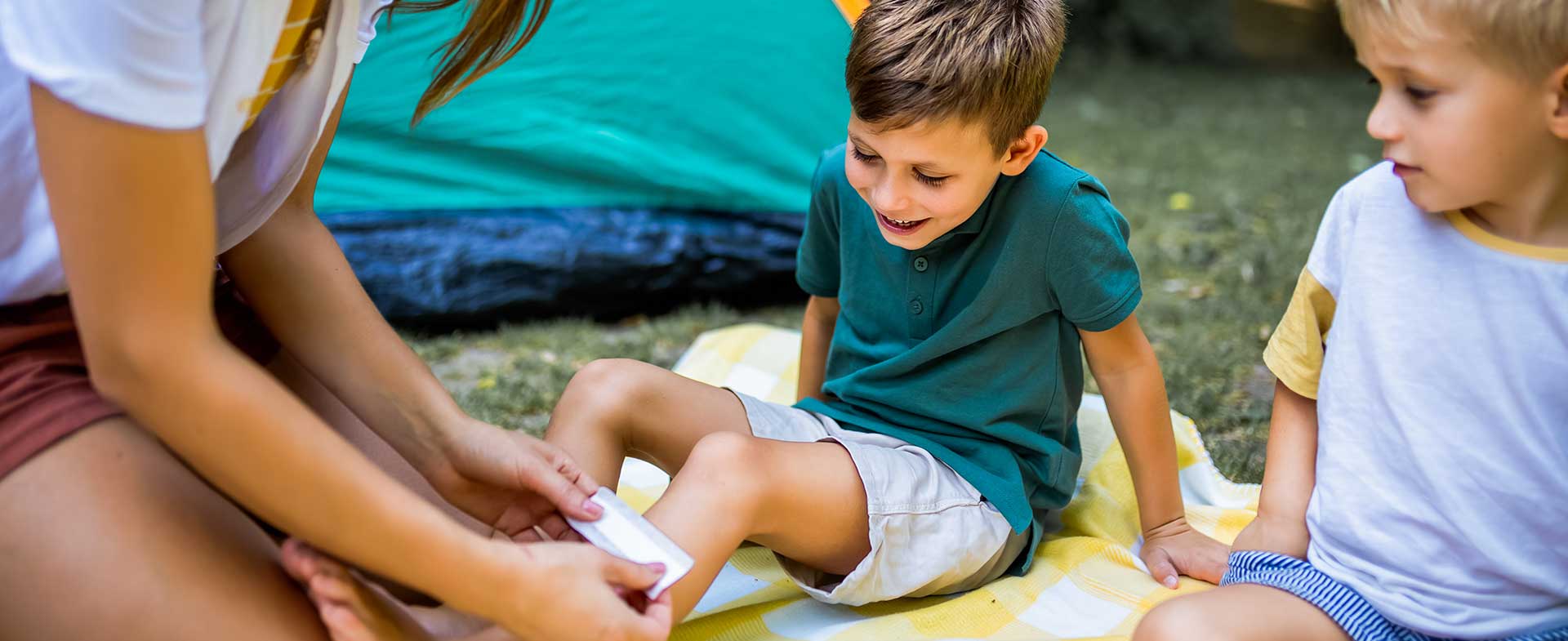 A young woman tends to a child's scraped knee. They are seated with another child on a picnic blanket near a green and orange tent in a grassy area. summer camp ailments
