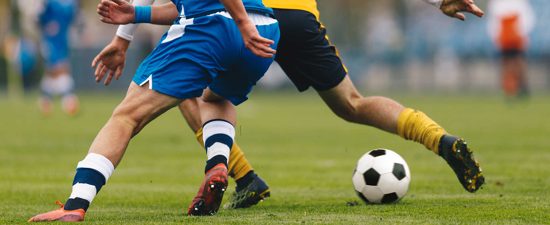 Two men playing soccer on turf in uniforms. Photo shows closeup of their legs colliding with the soccer ball. dummy desktop Image