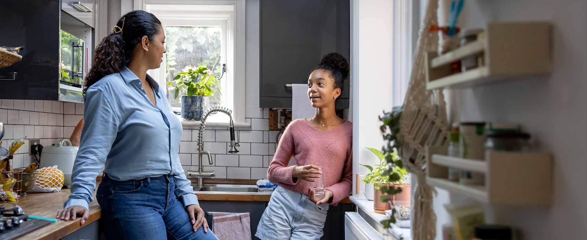 A mother and a teen daughter are chatting in a modern kitchen. They are casually dressed. The kitchen has wooden countertops, potted plants and shelves with various items. air traffic controller parents