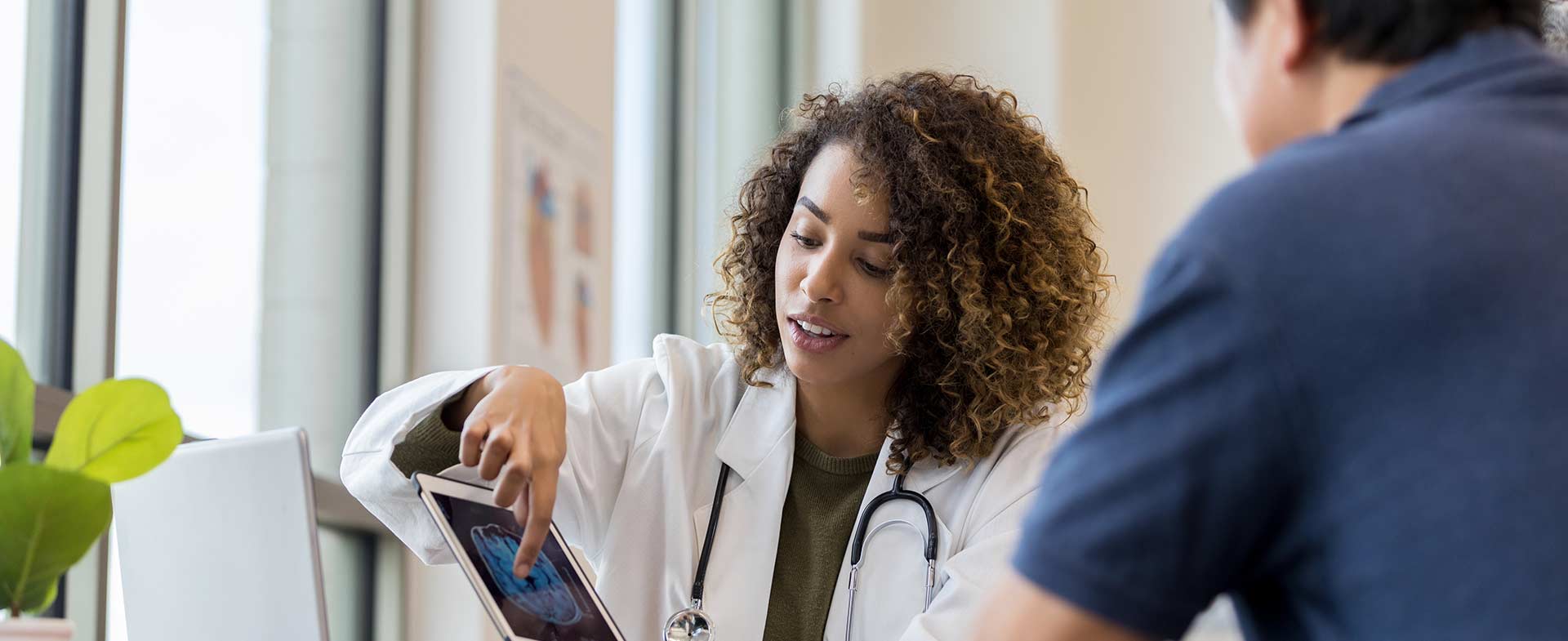 A female medical professional in a white coat shows a patient a brain scan on a tablet screen, sitting in a well-lit room with large windows. CAA and dementia risk