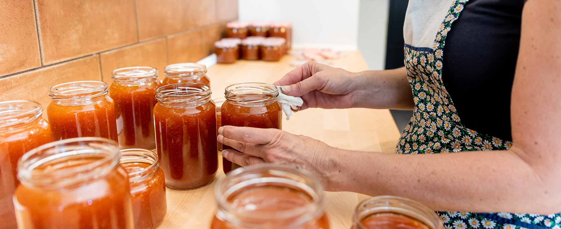 A woman carefully sealing jars of orange jam on a wooden countertop. Multiple filled jars are aligned in the background, and she is wearing an apron with a floral pattern. canning safety