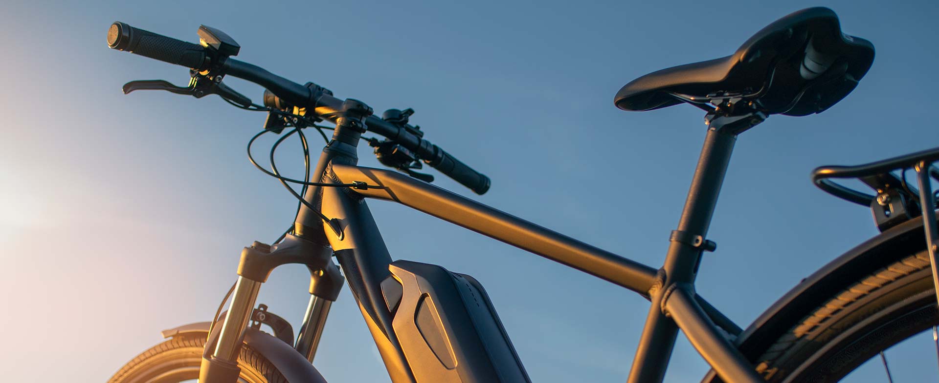 A sleek black electric bicycle with a prominent battery pack is set against a clear blue sky in the background, captured in warm sunlight. ebikes