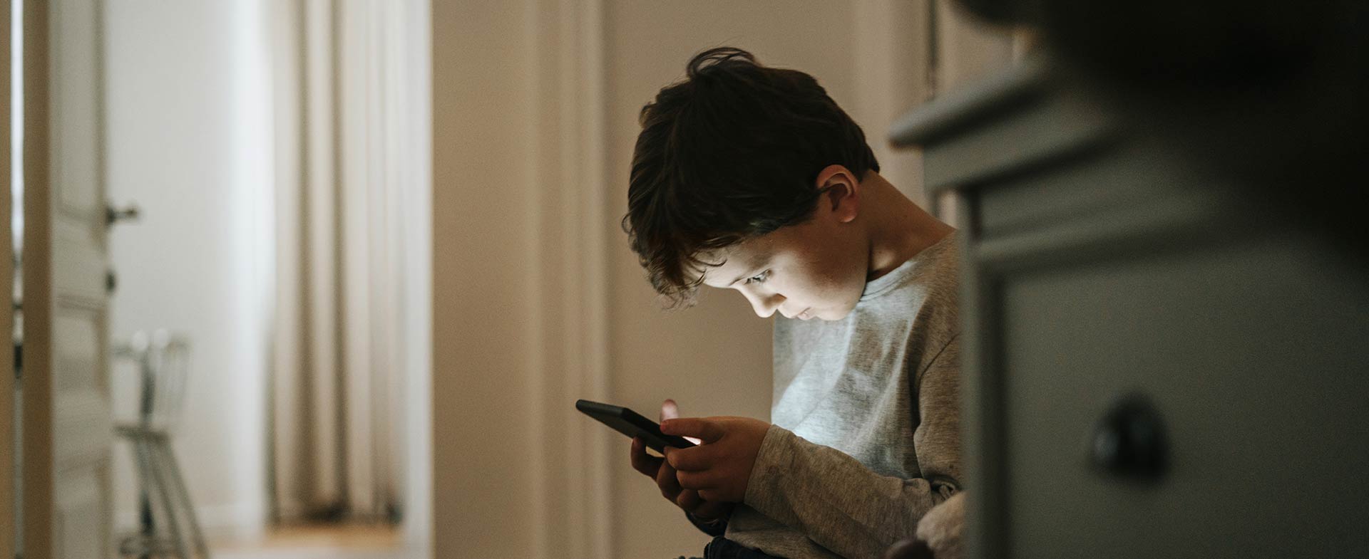 A young boy sitting alone in a soft lit room indoors, quietly focused on a smartphone in his hands. kids and cellphones