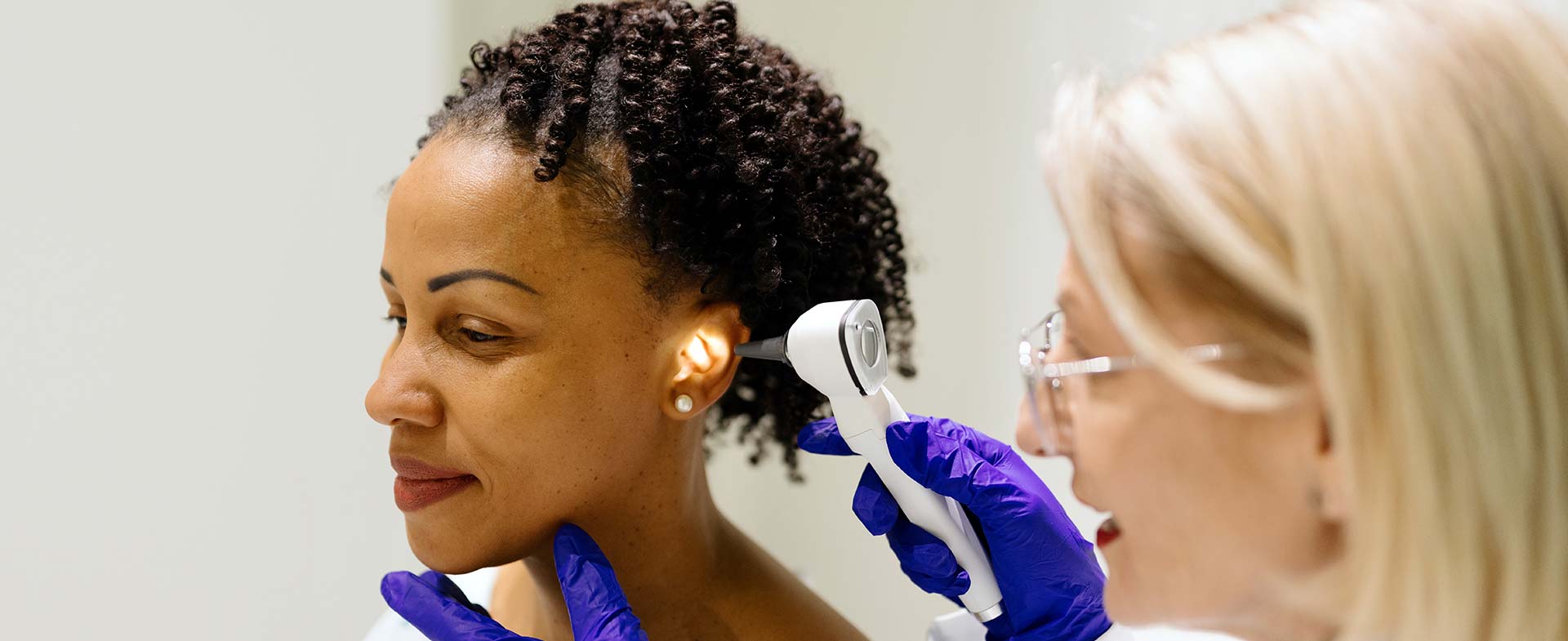 A woman is seated having an ear examination by a female healthcare professional using an otoscope. gene therapy for otof hearing loss