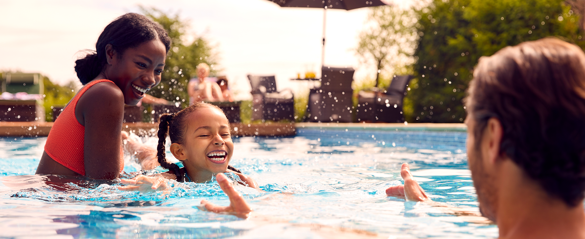 A family of three swimming outside in a pool. The mom is wearing a neon pink two-piece bathing suit and smiling, the daughter has braids in her hair and is smiling and the dad has his arms opened out to his smiling child. dummy desktop Image
