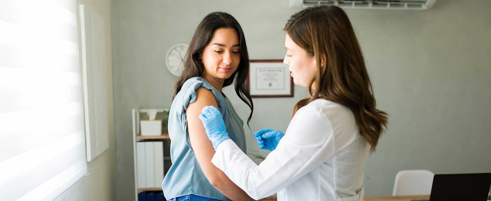 Woman with dark hair sits in doctor's exam room as a female provider prepares to administer a vaccine. flu vaccine 2026