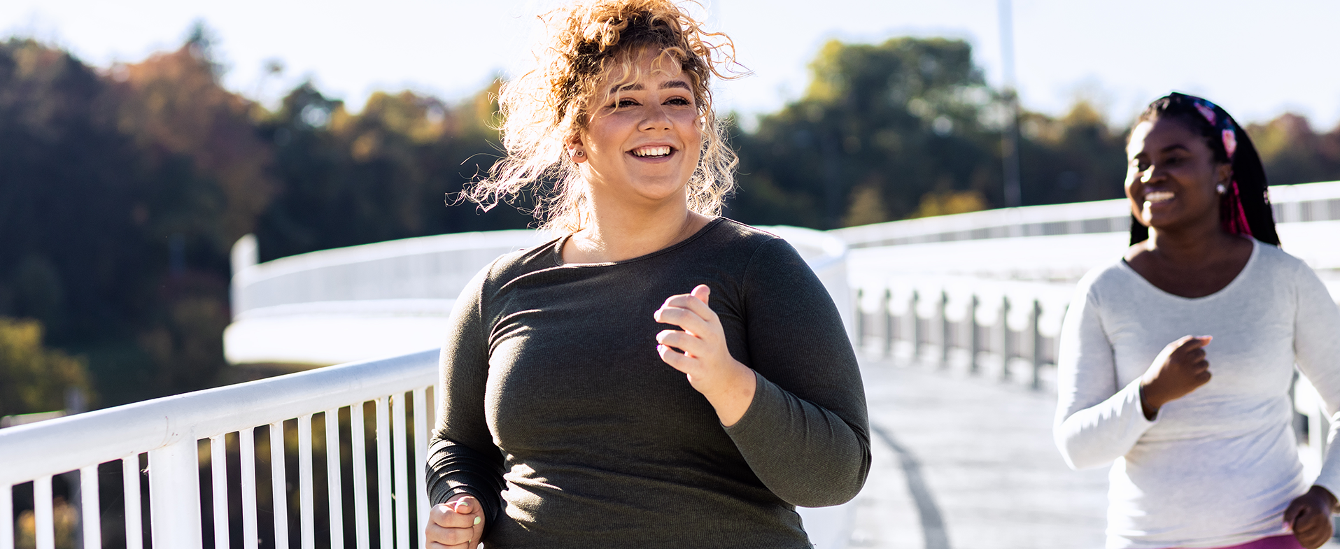 Two friends running outside. One is wearing a black long-sleeved running shirt and multi-colored patterned leggings. The other is wearing a white long-sleeved running shirt and hot pink leggings. Both are smiling. dummy desktop Image