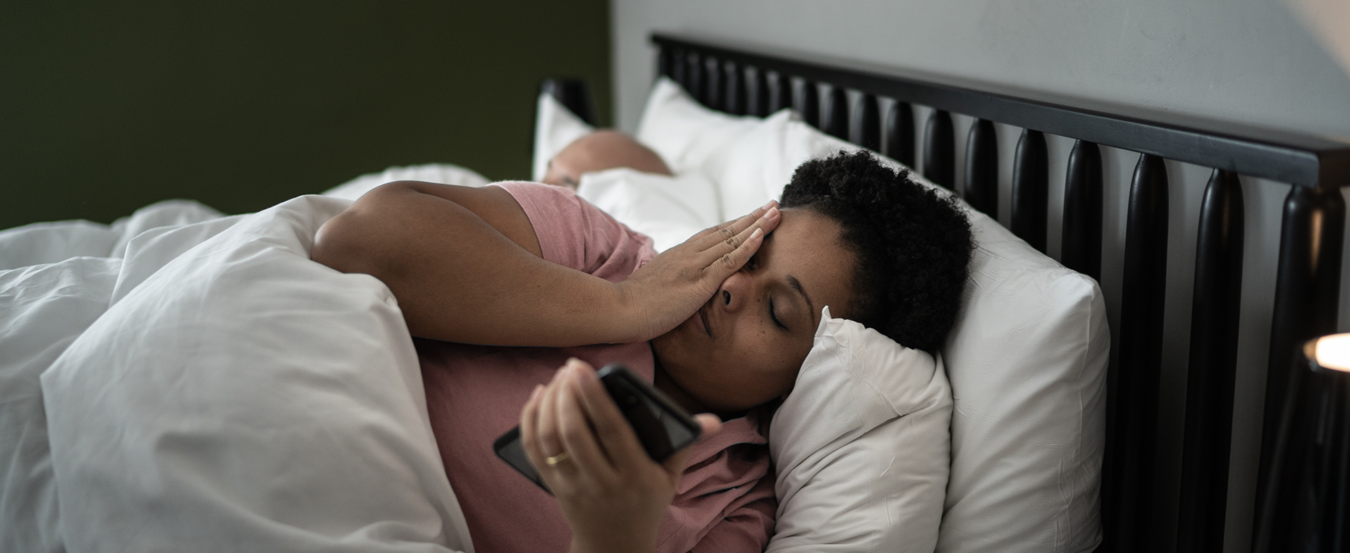 Woman with short curly hair laying in bed wearing a rose-colored t-shirt. She has her hand over her face and is holding her phone. dummy desktop Image