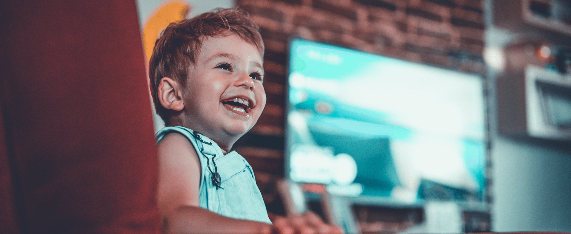Toddler boy smiling wearing light green overalls. He is sitting on a chair and there is a TV on in the background. dummy desktop Image
