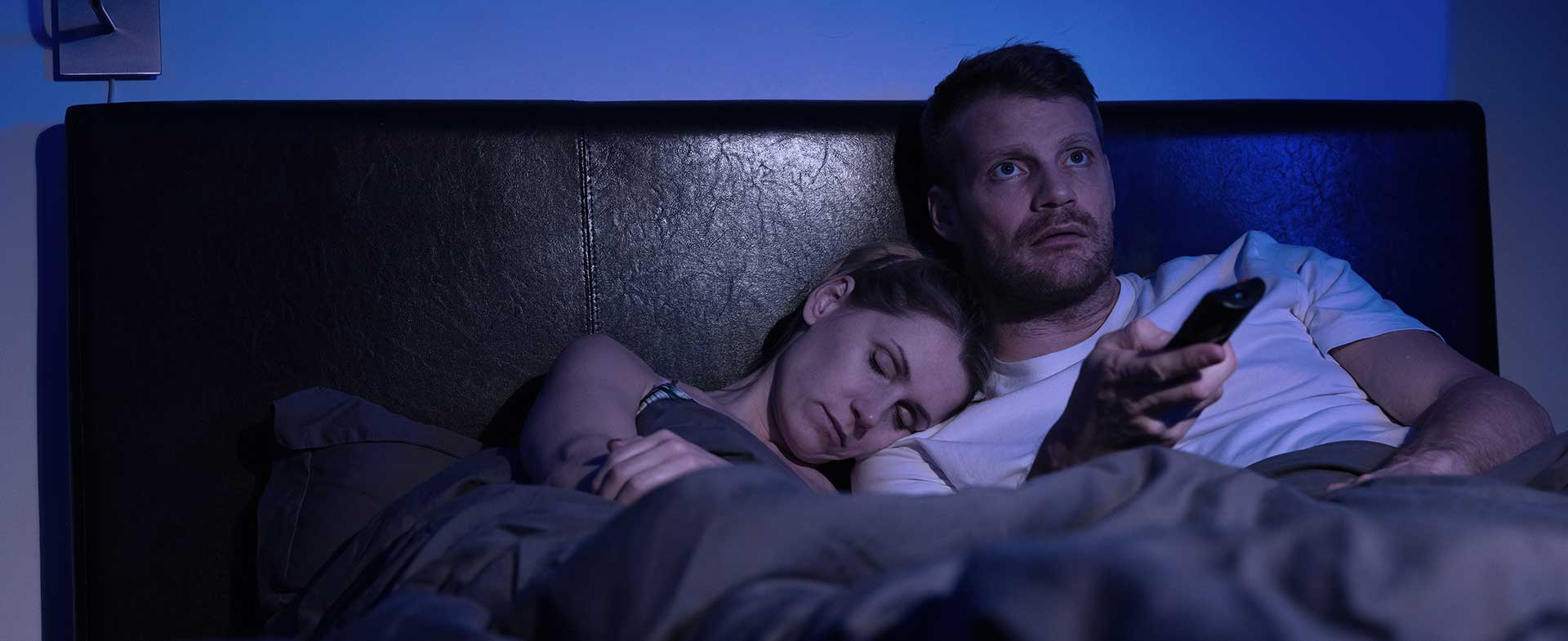 A couple sits in bed at night. The woman is asleep on the man's shoulder while he stays awake and changes the TV channel. early bird vs night owl heart health