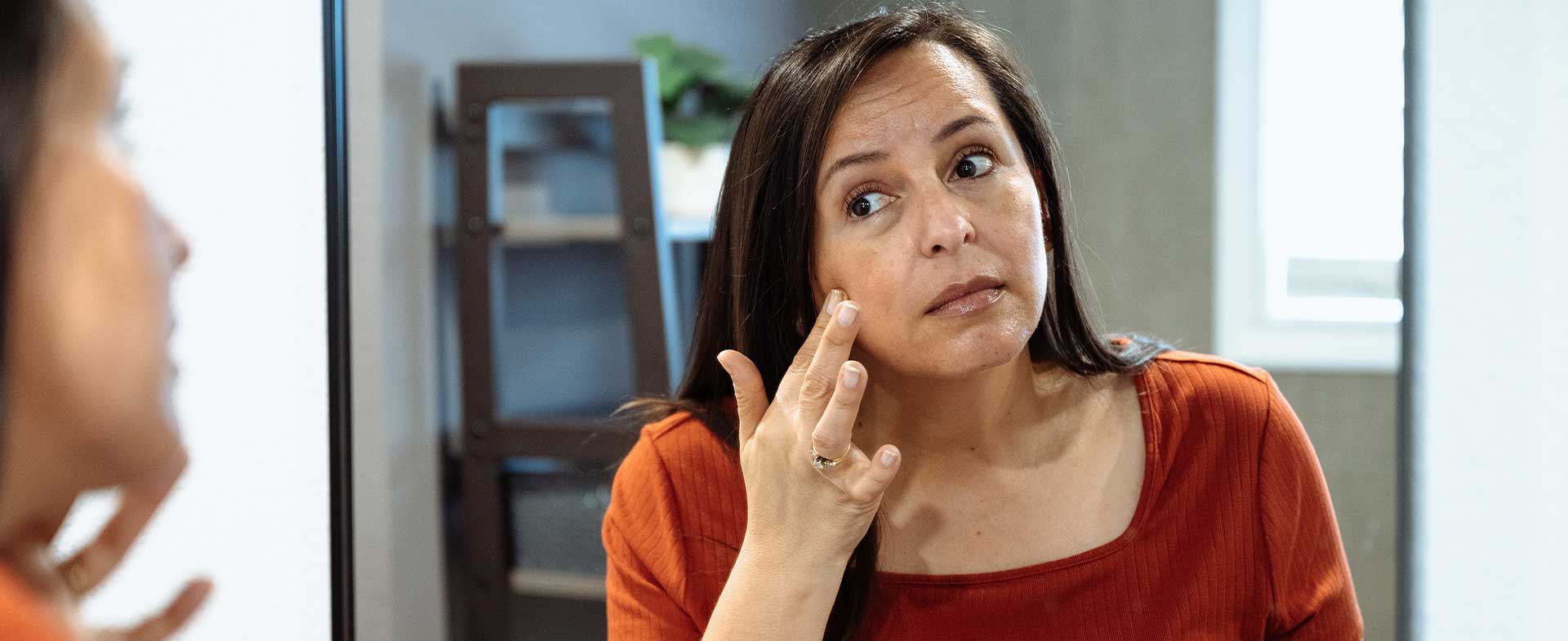 A middle aged woman looks at herself in the bathroom mirror with a hand on her face. ozempic face