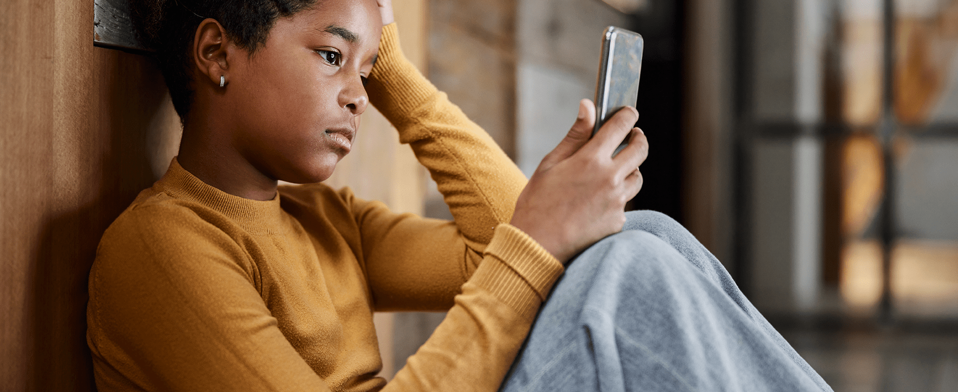 Girl sitting down at school in the hallway with a sad expression on her face and reading something on her phone. She is wearing a mustard yellow long-sleeve, crewneck sweater and blue jeans. bullying awareness