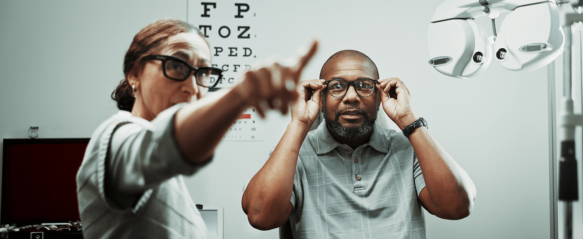 Older man getting an eye exam. He is wearing a gray polo T-shirt and holding glasses up to his eyes while a doctor points at something for him to see. dummy desktop Image