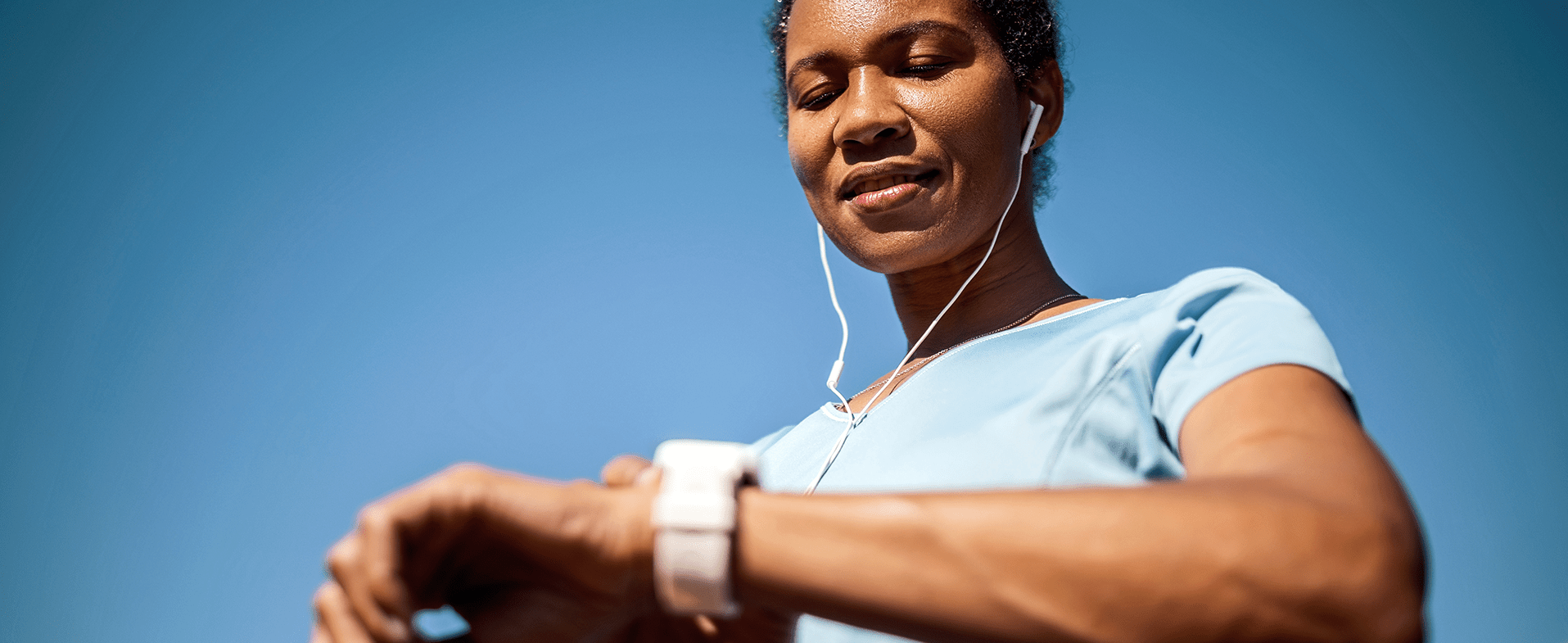 Woman wearing a light blue t-shirt standing outside on a blue-sky day looking at her smart watch. woman looking at smart watch