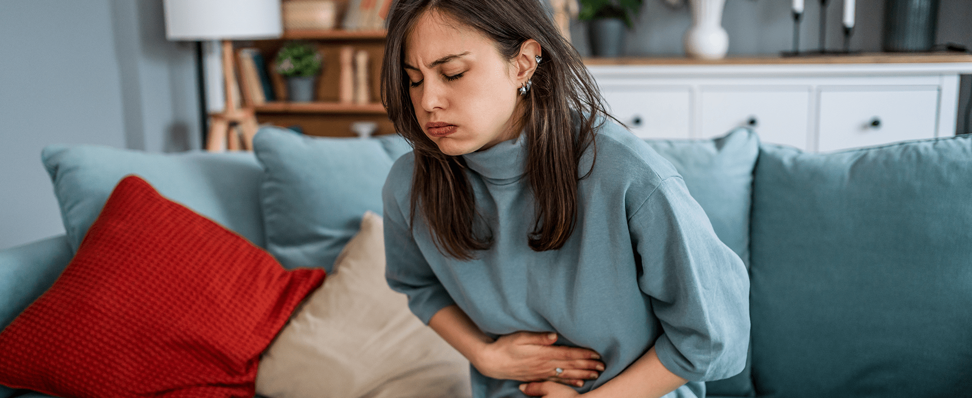Woman with dark brown long hair wearing a blue turtleneck sweater. She is sitting on the couch and has her hands on her stomach. dummy desktop Image