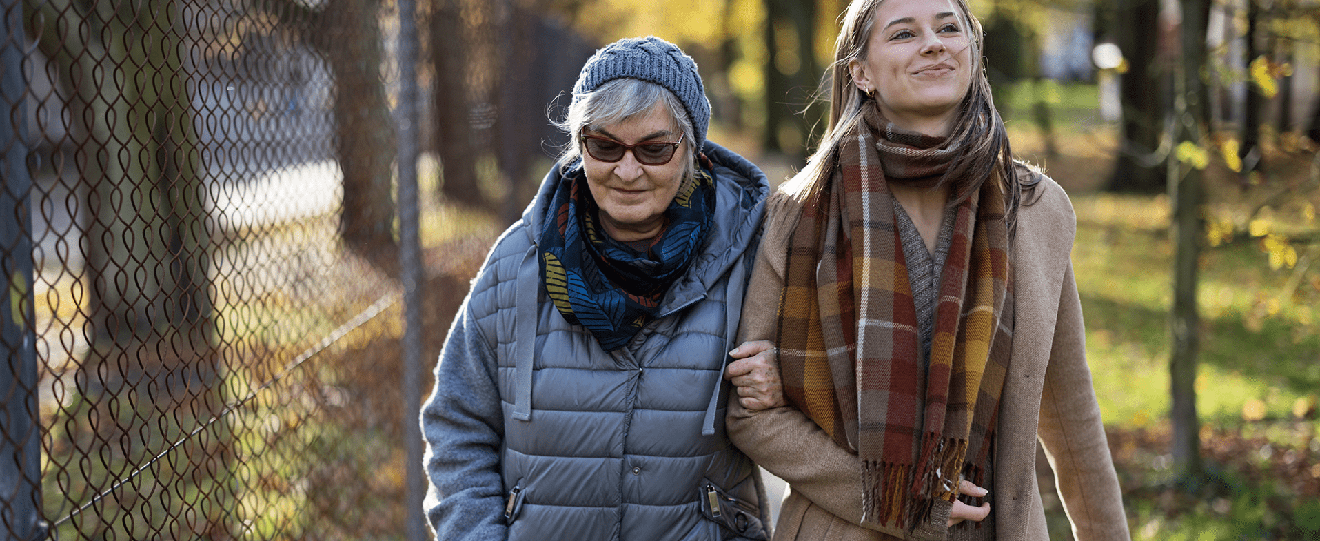 Older woman with gray short hair wearing a blue hat, gray puffer coat, teal scarf and sunglasses. She is walking outside with a younger woman who has blonde hair and is wearing a camel coat and a multi-colored plaid scarf. dummy desktop Image