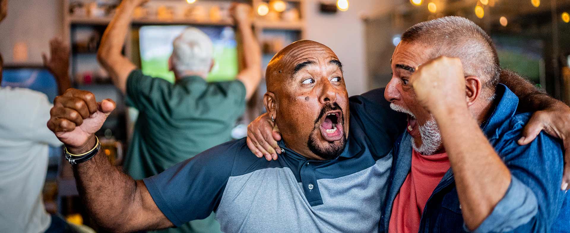 A group of people excitedly celebrating in a room, likely due to a sports event on a TV in the background. Two people in the foreground are embracing with joyful expressions, surrounded by warm lighting and hanging bulbs. sporting event heart attack