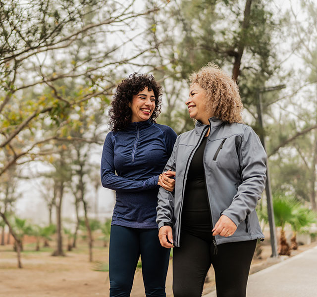Two women walking and smiling at each other in a tree-lined park path. They are dressed in casual athletic wear. The background features trees and an overcast sky. 10k to 7k steps