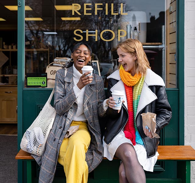 Two women sitting on a bench outside a "Refill Shop," holding coffee cups and smiling. One wears a checked coat and yellow pants, and the other wears a black and white coat with a colorful scarf. The shop door is open, with visible shelves and products inside. rx for laughter