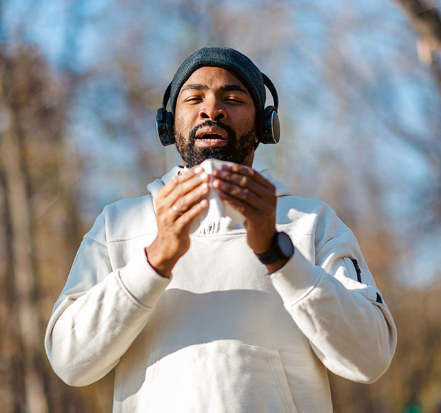 A man standing outdoors in a forested area holding a tissue, ready to sneeze. They are wearing headphones, a light-colored hoodie and a beanie. The background shows bare trees and a clear blue sky, suggesting a cool day. working out sick