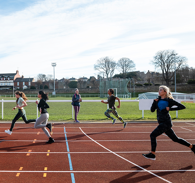 High schoolers wearing leggings, athletic tops and sneakers and running on a track outside. One person is standing alongside the track watching. people running on track