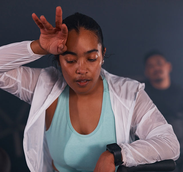 A woman wearing a light blue top and white jacket wipes her forehead after working out energetically. Another person is visible in the background, slightly out of focus. The setting appears dimly lit, creating a dynamic atmosphere. hot workouts