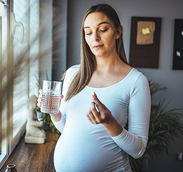 A pregnant woman with long hair and a long sleeve white maternity shirt holds a glass of water and a white pill. She stands inside near a window with bright natural light. aspirin use during pregnancy