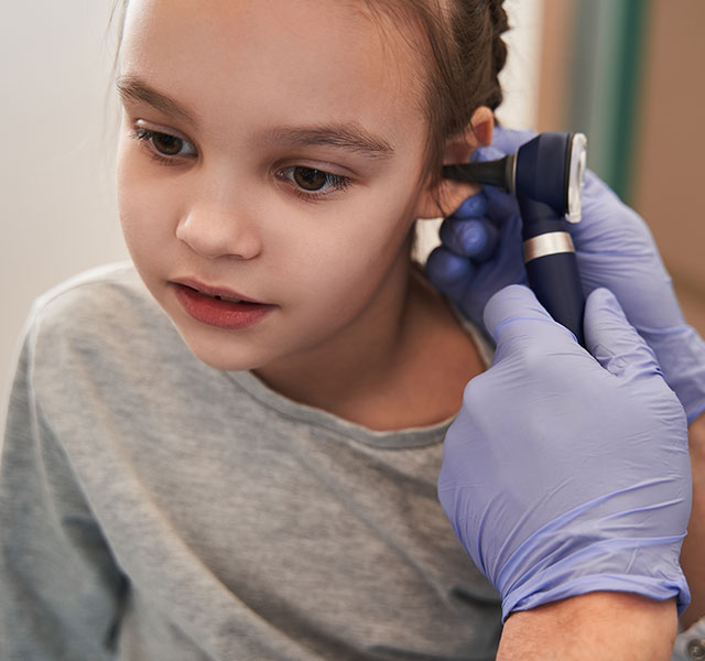 A person wearing blue gloves examines a young girl's ear with an otoscope in a medical setting. kid ear infections