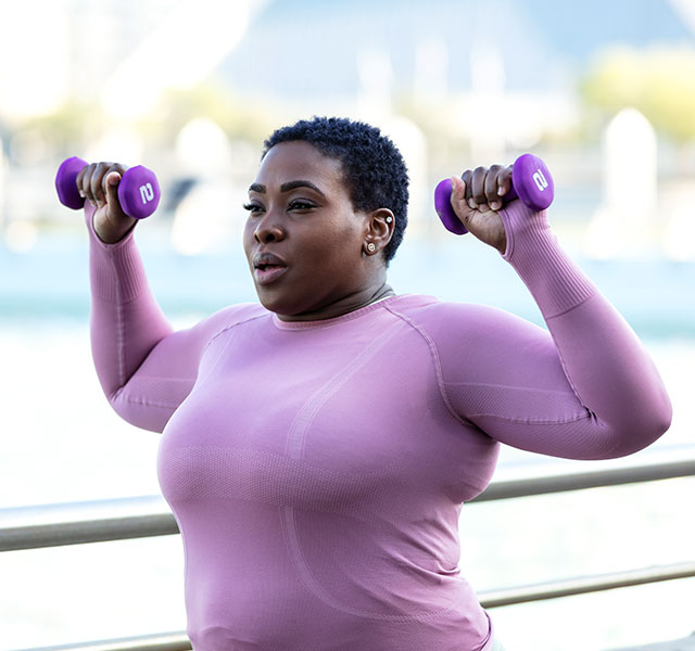 A Black woman in a purple long-sleeve shirt exercises outdoors with lightweight purple dumbbells. The background includes a blurred view of water and buildings. reversing type 2 diabetes