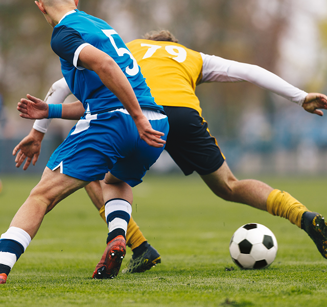 Two men playing soccer on turf in uniforms. Photo shows closeup of their legs colliding with the soccer ball. dummy mobile image