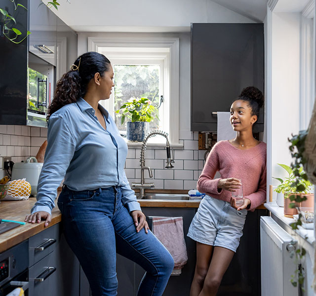 A mother and a teen daughter are chatting in a modern kitchen. They are casually dressed. The kitchen has wooden countertops, potted plants and shelves with various items. air traffic controller parents