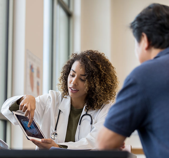 A female medical professional in a white coat shows a patient a brain scan on a tablet screen, sitting in a well-lit room with large windows. CAA and dementia risk
