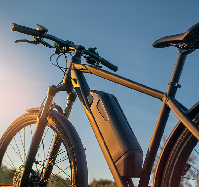 A sleek black electric bicycle with a prominent battery pack is set against a clear blue sky in the background, captured in warm sunlight. ebikes