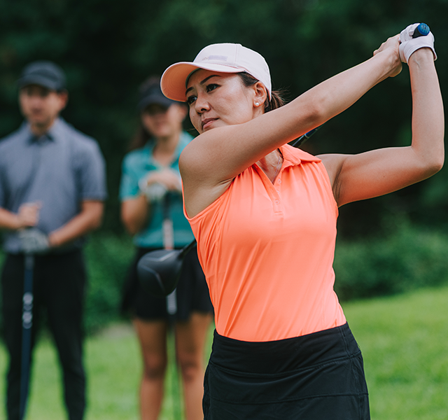 Female athlete outside playing golf. She is wearing a white visor on her head and a neon orange sleeveless, collared athletic shirt. Two golfers - a male and female - are standing in the background watching her. dummy mobile image