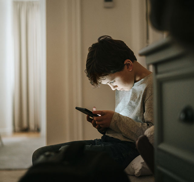 A young boy sitting alone in a soft lit room indoors, quietly focused on a smartphone in his hands. kids and cellphones