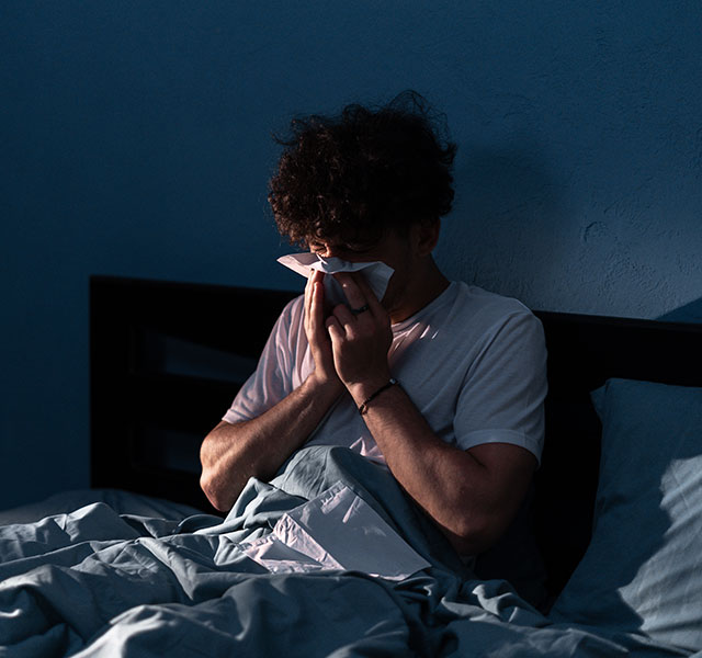 A person sitting up in bed in a dimly lit room, using a tissue to blow their nose. The bedding is a muted blue color. nighttime congestion