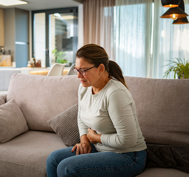 A person sitting on a sofa in a modern living room, holding their stomach and appearing uncomfortable. cyclosporiasis outbreak
